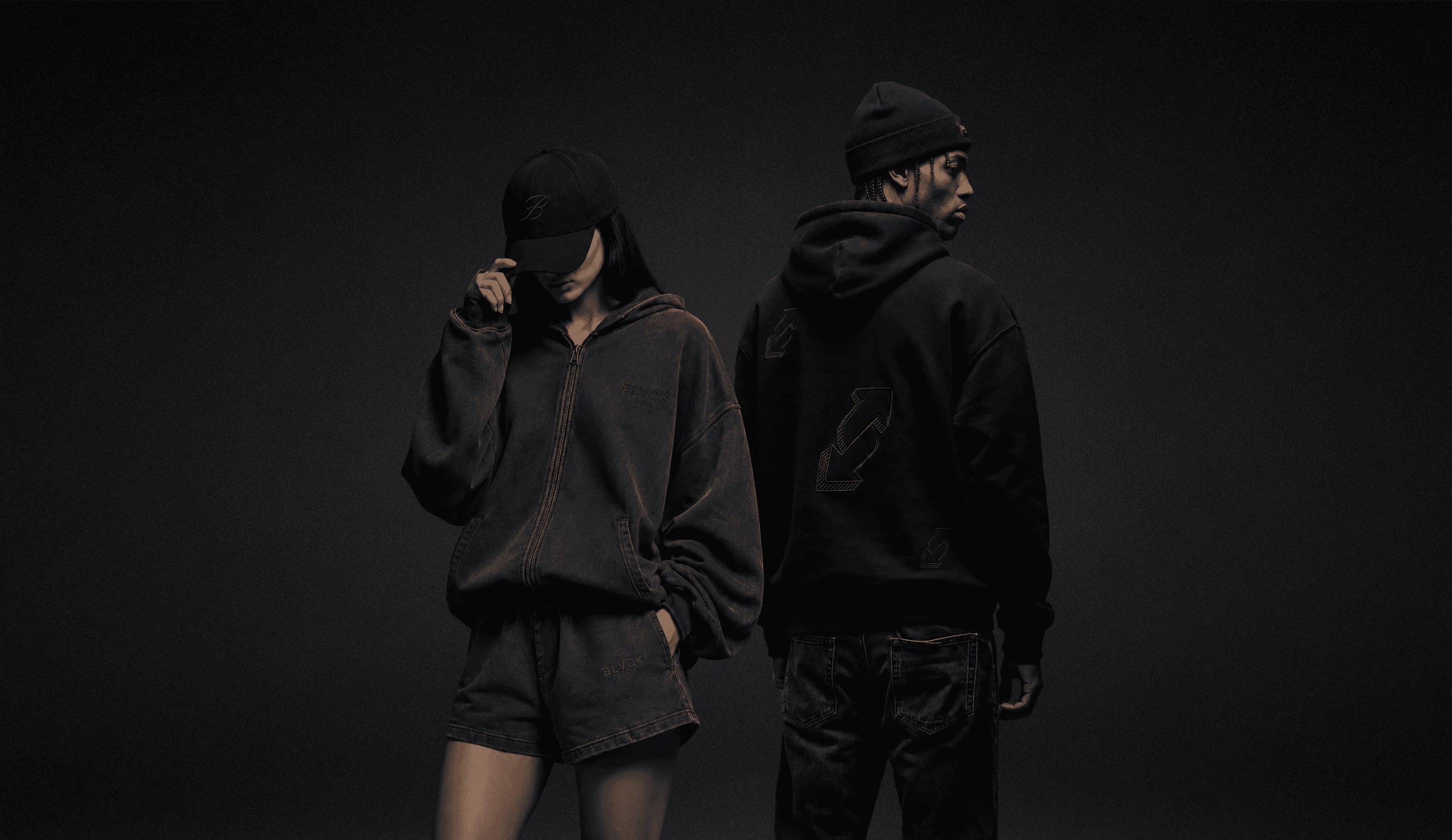 Two models wearing all-black BLVCK hoodies and accessories